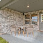 A covered patio area with a small round table and two wicker chairs on a concrete floor, attached to a brick house, with green grass and a wooden fence visible outside.