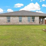 A single-story brick house with four windows and a covered patio, set in a large, neatly mowed backyard with a wooden fence and a small landscaped garden in one corner under a blue sky with white clouds.