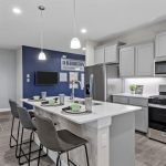 Modern kitchen with gray cabinets, stainless steel appliances, and a white island with three gray barstools. In the background, there’s a dining table with green chairs and a blue accent wall.