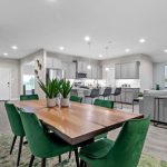 Modern open-concept dining and living area with a wooden table, six green velvet chairs, potted plants, and a sofa facing a wall-mounted TV. The kitchen with gray cabinets and bar stools is visible in the background.