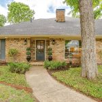 A single-story brick house with a gray shingle roof, large windows with blue shutters, a central front door flanked by potted plants, and a curved walkway through a landscaped front yard with trees and shrubs.