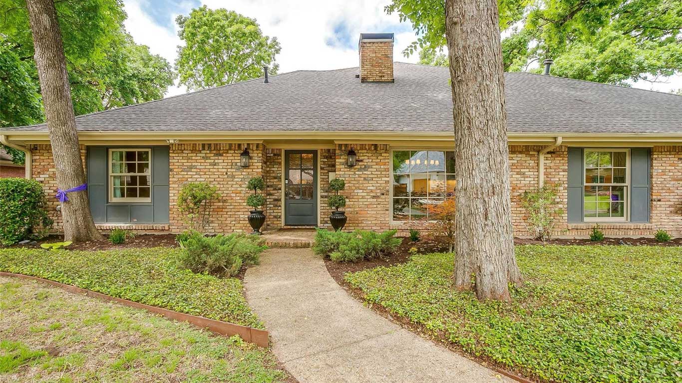 A single-story brick house with a gray shingle roof, large windows with blue shutters, a central front door flanked by potted plants, and a curved walkway through a landscaped front yard with trees and shrubs.