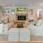 A bright, spacious living room with white sofas, a stone fireplace, a TV above the mantel, built-in shelves, light wood floors, and a view into an adjoining kitchen and hallway with a staircase.