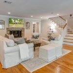 A spacious living room with hardwood floors, white sofas, a patterned rug, a fireplace with a TV above, built-in shelves, and a staircase leading upstairs. Family photos and decor accents add a cozy touch.