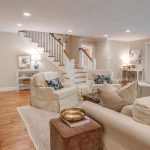 A spacious, light-filled living room with beige sofas, armchairs, and throw pillows, a fireplace, a TV mounted above, wood floors, a staircase, and large glass doors leading to a green outdoor area.