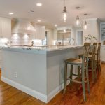 A modern kitchen with white cabinets, marble countertops, pendant lights, and four wooden bar stools at a large island. The kitchen opens to a cozy living area with built-in shelves and framed photos.