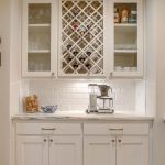 A built-in home bar area with white cabinets, a wine rack holding bottles, glassware on shelves, a coffee machine on the countertop, jars filled with corks, and white subway tile backsplash.