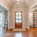 A bright entryway with a wooden front door, hardwood floors, light-colored walls, and a geometric ceiling light. To the right is a built-in bookshelf, and to the left is a living room with large windows and white curtains.