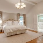 A spacious, elegant bedroom with a large bed, white bedding, two windows with floral curtains, a chandelier, armchair, side tables with lamps, and an open doorway leading to a bright bathroom.