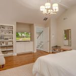 A spacious bedroom with a white bed, armchair, chandelier, built-in shelves, hardwood floors, and an open walk-in closet. Neutral-toned décor and a window provide a bright, cozy atmosphere.