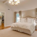 A spacious, well-lit bedroom features a large bed with white bedding, two bedside tables with lamps, floral curtains, a chandelier, a bench with a basket, and a walk-in closet visible through an open door.