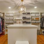 A spacious walk-in closet with white shelves and racks holding neatly organized clothes, shoes, and boxes. There is a central white island, wooden floor, and a geometric pendant light overhead.
