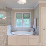 Modern bathroom with a glass-enclosed shower, a built-in oval bathtub below two windows with green shades, beige walls, and white cabinetry. Towel hangs on a rack next to the tub. Marble tile surrounds the bath area.