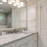 A modern bathroom with a marble countertop, built-in sink, white cabinets, a large mirror, and overhead lights. A glass shower with tile walls is visible in the background.