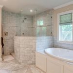 Elegant bathroom with a large glass-enclosed shower, white marble tile, a spacious built-in bathtub, double windows with green roman shades, and cream-colored cabinets and walls.