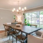 A bright dining room with a large wooden table, six mixed chairs, a chandelier, a light rug, and a sideboard. Big windows with white curtains provide a view of a green yard and houses outside. A leafy plant sits by the window.