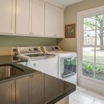 A laundry room with a black countertop, sink, white cabinets, a washer and dryer, and a large window showing a view of a suburban street and greenery outside.