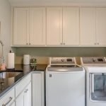 A laundry room with white cabinets, a stainless steel sink with a pull-down faucet, a washing machine, a dryer, paper towels, and framed botanical prints on light green walls. A window lets in natural light.