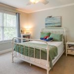 A bright bedroom with a white metal bed frame, green bedding, and a plush toy on the bed. There is a large window with blue curtains, a dresser, a nightstand, and a beach-themed painting above the bed.
