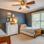 A cozy bedroom with two twin beds, navy blue walls, a ceiling fan, large window with blinds, a white dresser with framed art above it, and a bookshelf filled with books.