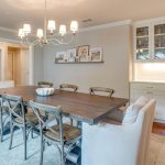 A bright dining room with a wooden table, eight chairs, a chandelier, white cabinets with glass doors, and decorative items on a shelf. Hardwood floors and a large rug complete the cozy, modern space.