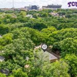 Aerial view of a tree-filled residential neighborhood with a large stadium in the background. A house is highlighted with an icon, and the TCU logo appears in the upper right corner.