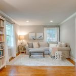 A cozy living room with light gray walls, a beige sectional sofa, a tufted bench, a white area rug, a side table with a lamp, two artwork pieces, and bookshelves filled with books on both sides.