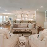 A spacious, open-concept living room with a beige sofa, two armchairs, a tufted coffee table, and a fireplace leads into a modern kitchen with white cabinets, barstools, and pendant lights. Large windows provide natural light.