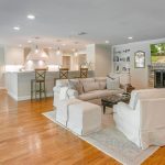 Spacious open-concept living area with hardwood floors, white furniture, a fireplace with a TV above it, and a kitchen with barstools. A dining table sits to the left near large windows. The room is bright and inviting.