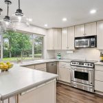 Modern kitchen with stainless steel appliances, light-colored cabinets, an island with a bowl of lemons, pendant lighting, large window overlooking a green yard, and wood flooring.