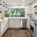 Modern kitchen with white cabinets, stainless steel appliances, a large window, pendant lights, and dark wood flooring. A bowl of lemons sits on the counter, and greenery is visible outside the window.