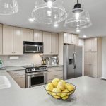 Modern kitchen with light wood cabinets, stainless steel appliances, and a gray countertop. A bowl of lemons sits on the island, and pendant lights hang above. There is a sink by a window on the left.