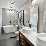 Modern bathroom with gray tiled walls, freestanding white bathtub, glass-enclosed shower, double vessel sinks on a marble countertop, large mirrors, and hexagon floor tiles. Contemporary fixtures and natural light.