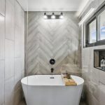 Modern bathroom with a freestanding white tub, herringbone tile accent wall, black fixtures, a window, built-in niche with bath products, and a wall-mounted showerhead. A wooden tray sits on the tub.