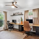 A bright home office with two desks, computers, and chairs facing windows with beige curtains. A ceiling fan, indoor plant, framed pictures, and a rug complete the modern, organized workspace.