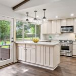 Modern kitchen with light wood cabinets, stainless steel appliances, a marble countertop island with a bowl of lemons, pendant lights above, and large windows overlooking a green backyard. French doors lead outside.