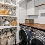A modern laundry room with front-load washer and dryer, wooden countertop, white cabinets, open shelves with baskets and mirrors, and black-and-white floral wallpaper accent. Laundry hampers and decor complete the space.