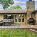 A cozy backyard patio with outdoor wicker furniture, a small table, a barbecue grill, string lights, and French doors, set against a brick house with green grass and trees in the background.