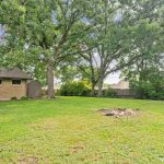 A spacious backyard with green grass, large shade trees, a small fire pit, a patio seating area, and a brick house with a shed. A chain-link fence runs along one side of the yard.