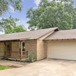 Single-story brick house with a front porch, two-car garage, and a concrete driveway. The yard has some shrubs and mature trees, with a wooden fence and green foliage in the background under a partly cloudy sky.