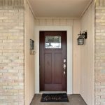 A brown wooden front door with a glass panel at the top, surrounded by light brick walls. A black outdoor lantern and mail holder are mounted on the wall, and a doormat says, “It’s good to be home.”.