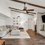 A modern living room with a gray sectional sofa, a white rug, and wall art. A large TV is mounted above a white brick fireplace. Exposed wooden ceiling beams, wood floors, potted plants, and neutral decor create a cozy, inviting space.