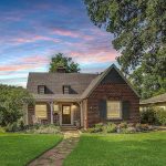 A charming brick house with a steep roof, large windows, and a front porch sits amid a well-kept lawn and garden, surrounded by mature trees, under a colorful evening sky.
