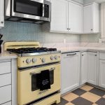 A kitchen with white cabinets, a vintage yellow gas stove with a towel hanging on the handle, a knife block, a microwave above the stove, checkered flooring, and a window with shutters above the sink.