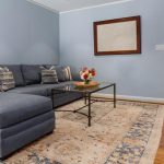 A cozy living room with a blue sectional sofa, decorative pillows, a glass coffee table with a flower arrangement, an ornate rug, wooden floors, and light blue walls with a framed artwork.