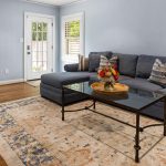 Living room with a blue sectional sofa, patterned pillows, a glass coffee table with a flower arrangement, a beige and blue rug, wooden floor, and light blue walls. A door and windows let in natural light.