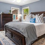 A cozy bedroom with blue-gray walls, dark wood furniture, a bed with gray and white bedding, decorative pillows, two windows with outdoor views, and a patterned rug on a wooden floor.