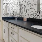 A modern bathroom with a black countertop, dual sinks, white cabinets, and a decorative black swirl wallpaper, with a glimpse of a bedroom featuring wood floors and a bed with hanging wreaths.