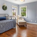 A bright, cozy room with hardwood floors, a metal daybed with blue bedding, an upholstered armchair with ottoman, green wreaths on the wall, and large windows letting in natural light.