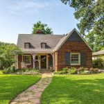 A charming brick house with dark shutters and a gabled roof sits behind a lush green lawn and garden, with a stone path leading to the front porch. Surrounded by trees and neighboring homes on a sunny day.